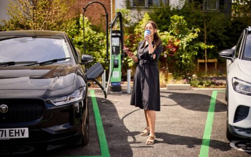 a a white woman in a black dress drinks a takeaway coffee while charging her EV